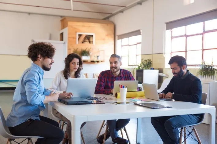 Dubai Business Setup - Four individuals working at a table with laptops and office supplies in a bright workspace.